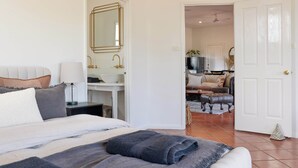 Ein Schlafzimmer mit Bett, Schreibtisch und Spiegel. Eine Tür führt zu einem Wohnbereich mit Sofa und Fernseher.