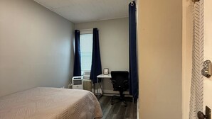 Quarto com cama, escrivaninha e cadeira. Há um radiador sob a escrivaninha e uma janela com cortinas.