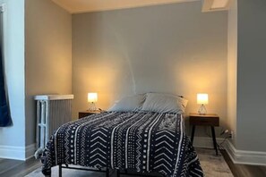 A bedroom with a bed, two bedside tables with lamps, a radiator, and a window.