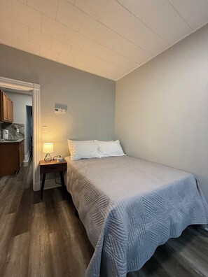 A bedroom with a bed, bedside table, lamp, and a view of the kitchen.