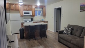 A compact kitchen with wooden cabinets, a white refrigerator, a microwave, a stove, and a sink. There are two bar stools at the kitchen island. A gray sofa is in the living area, and a door leads to another room.