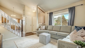 Un vaste salon avec un canapé gris, un pouf assorti et un fauteuil gris. Un escalier avec des rampes blanches mène à l’étage supérieur.