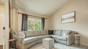 A living room with a sectional sofa, a small ottoman, a side table, and a framed picture on the wall. - Mid-ViewHouse Inn (Colorado Springs)