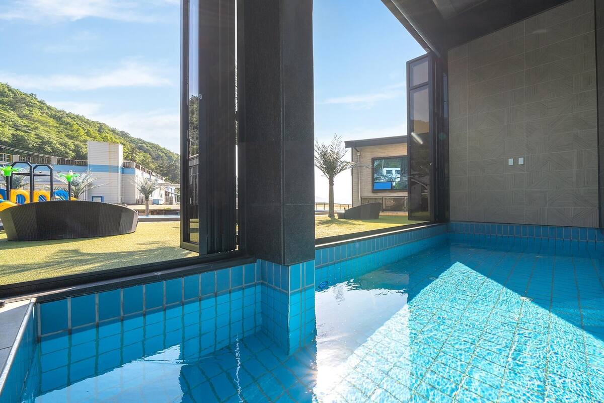 A modern indoor swimming pool with clear blue tiles, large windows, and a view of a playground and hills in the background.
