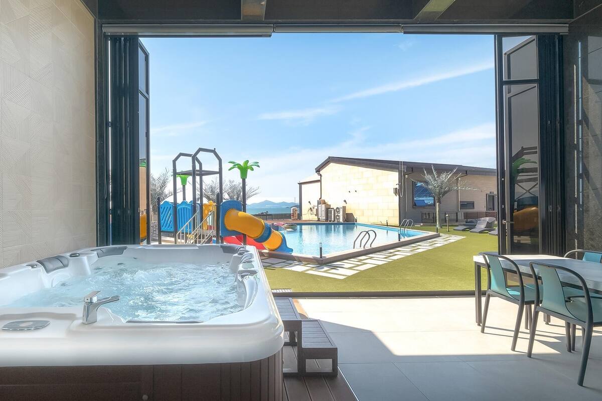 A modern outdoor pool area with a hot tub, a children's play structure, and a view of a building in the background.