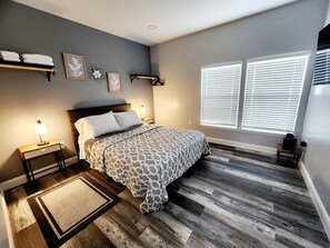 A bedroom with a bed, bedside lamps, a rug, a window with blinds, and a small table with a laptop.