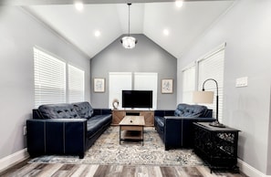 Un salon avec un plafond haut, un téléviseur à écran plat, deux canapés sombres, une table basse et un tapis à motifs.