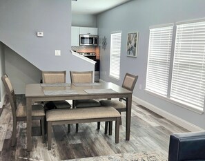 A dining area with a wooden table and chairs, a kitchen in the background, and a window with blinds.