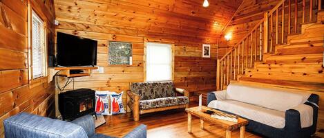 A cozy cabin interior with wooden walls, a television, a sofa, a chair, and a fireplace.