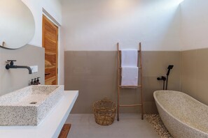 Un baño con un lavamanos rectangular, una bañera independiente, una puerta de madera, una cesta de mimbre y un perchero para toallas.