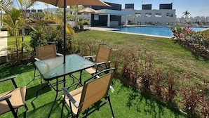 Un patio con mesa y sillas de vidrio, un paraguas grande y una piscina al fondo.