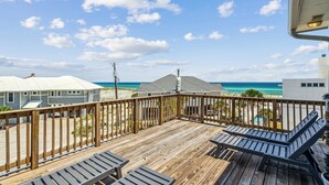 Une terrasse en bois avec des chaises longues, une vue sur la plage et des bâtiments en arrière-plan.