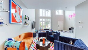 Un salon moderne avec un canapé, des fauteuils et une table basse. Un téléviseur est fixé au mur, et une enseigne au néon est visible.