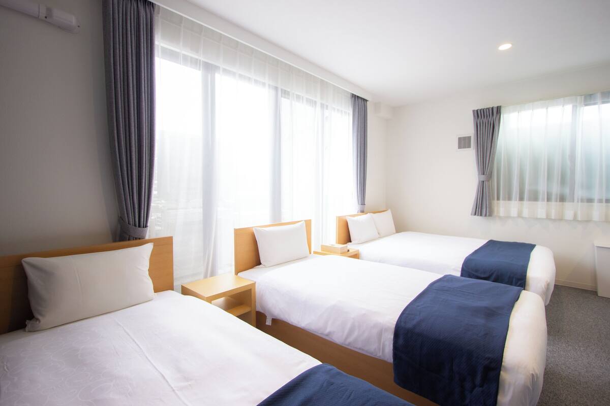 A hotel room with three beds, a wooden headboard, a nightstand, and a window with sheer curtains.