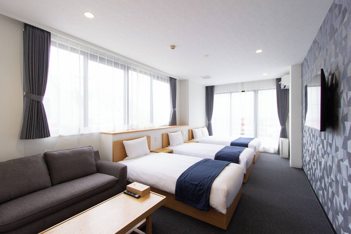 A hotel room with two beds, a sofa, a TV, and large windows.