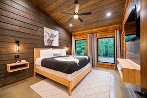 Une chambre à coucher située dans une cabane en bois, comprenant un lit, des tables de chevet, un ventilateur de plafond et une grande fenêtre avec des rideaux.