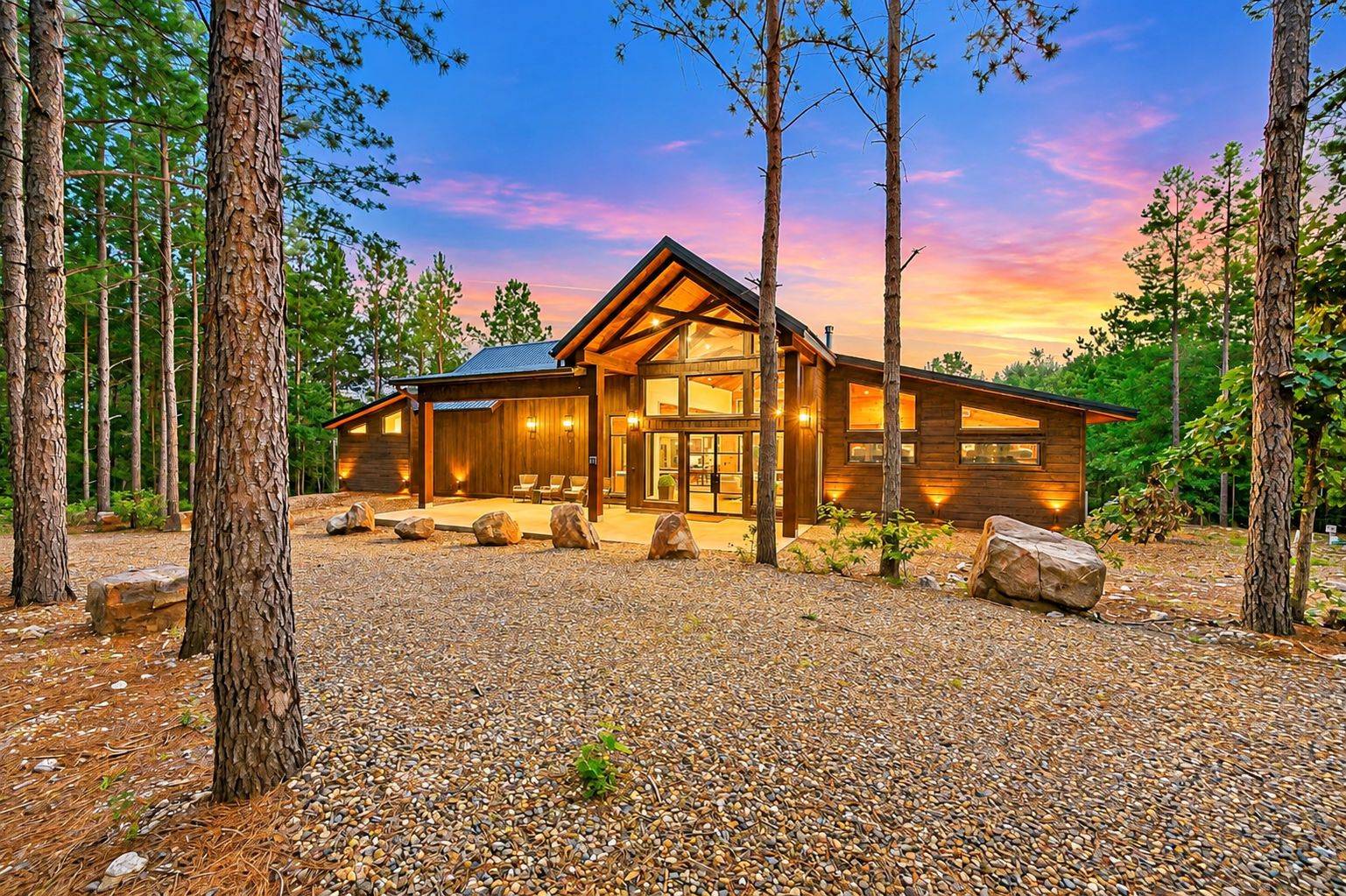 A modern log cabin with a large glass entrance, surrounded by a gravel driveway and large rocks.