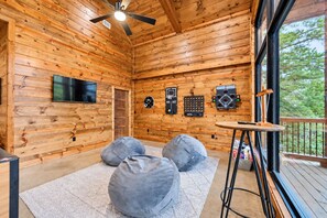 A wooden cabin interior with a ceiling fan, a television, a basketball hoop, and bean bags on a rug. - Spacious Luxe 4BR | Game Room | Fire Pit & Hot Tub (Broken Bow)