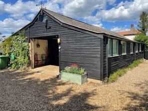 A black wooden building with a sloped roof, a visible door, and a small garden area with plants. - 2 Bed Barn - Sleeps 4 - Pets - Parking - Wifi (Downham Market)