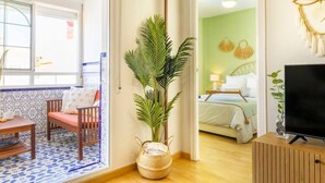 Een kamer met een houten vloer, een bed met wit beddengoed, een televisie op een houten dressoir, een plant en een raam met uitzicht.