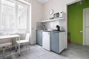 Una cocina compacta con un refrigerador blanco, un microondas y un lavamanos. Hay una mesa de comedor con una silla y una ventana con persianas.