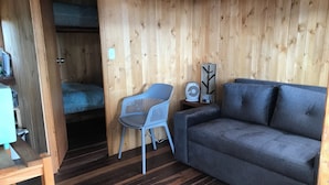 Quarto compacto com uma cama beliche, um sofá cinzento, uma cadeira azul e uma pequena mesa com um ventilador.