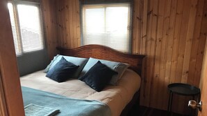 Quarto com painéis de madeira, uma cama, duas janelas com persianas e uma pequena mesa de cabeceira.