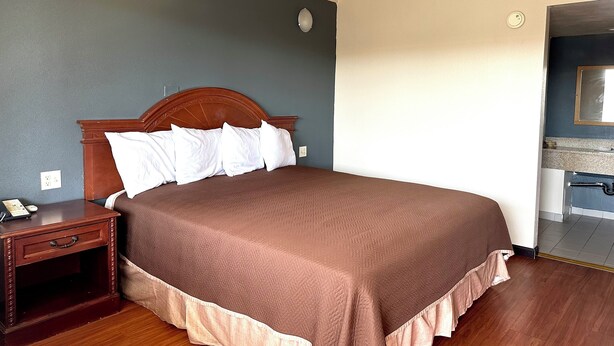 A neatly made bed with a wooden headboard, a bedside table with a lamp, and a wall-mounted light fixture. - Regency Inn Fresno (Fresno)