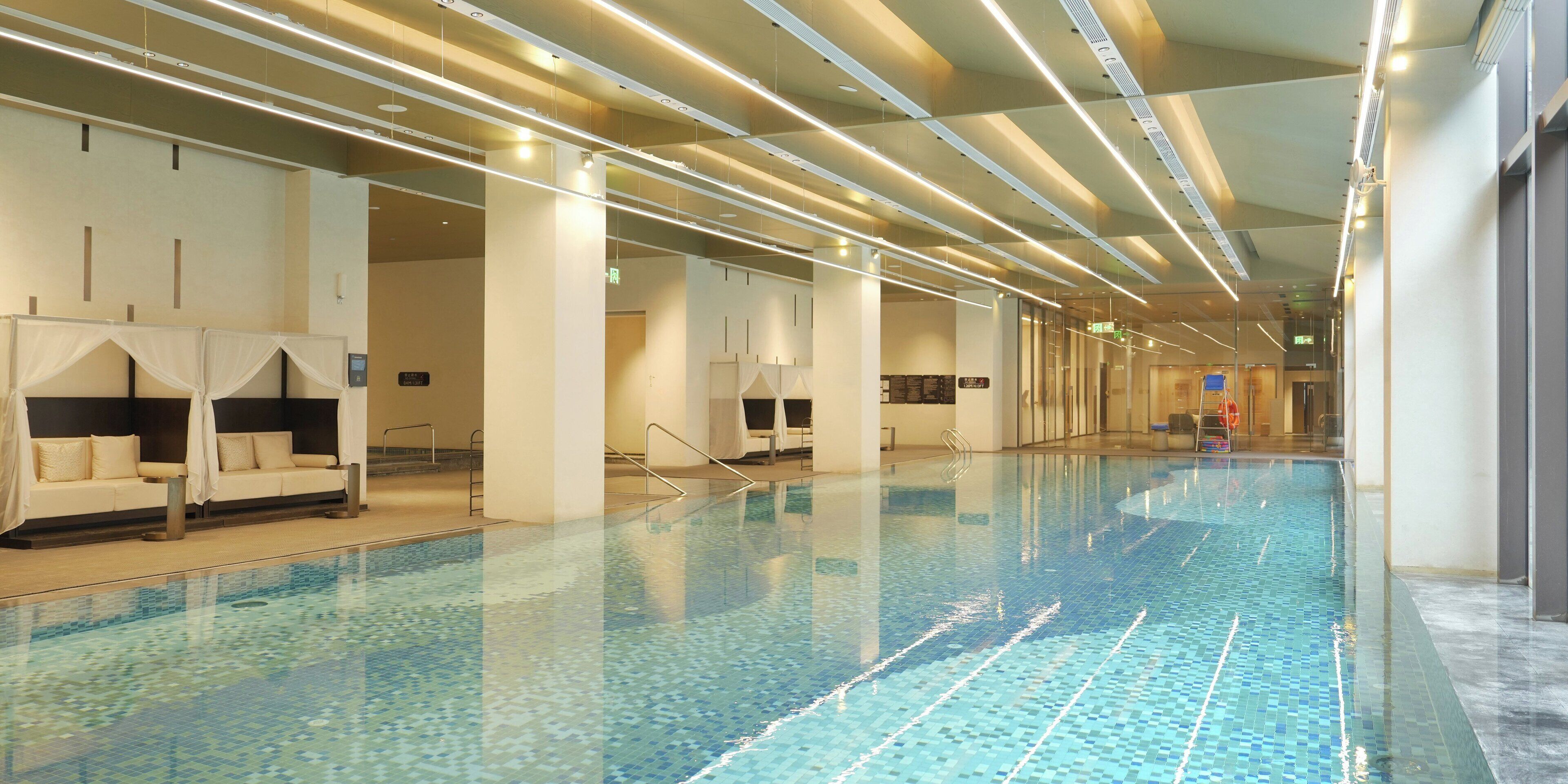 A long indoor swimming pool with clear water, surrounded by white walls and columns, and equipped with modern lighting fixtures.