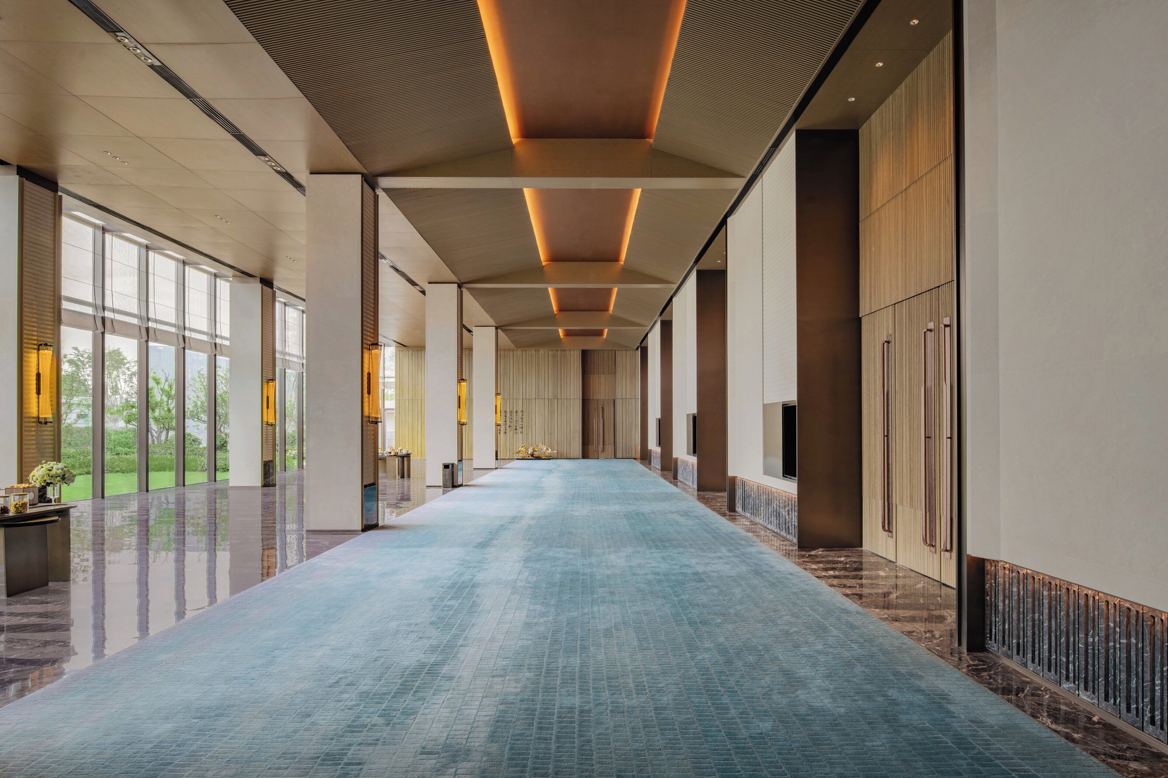 A long, narrow hallway with a blue carpet, large windows on one side, and a wooden panel wall on the other.