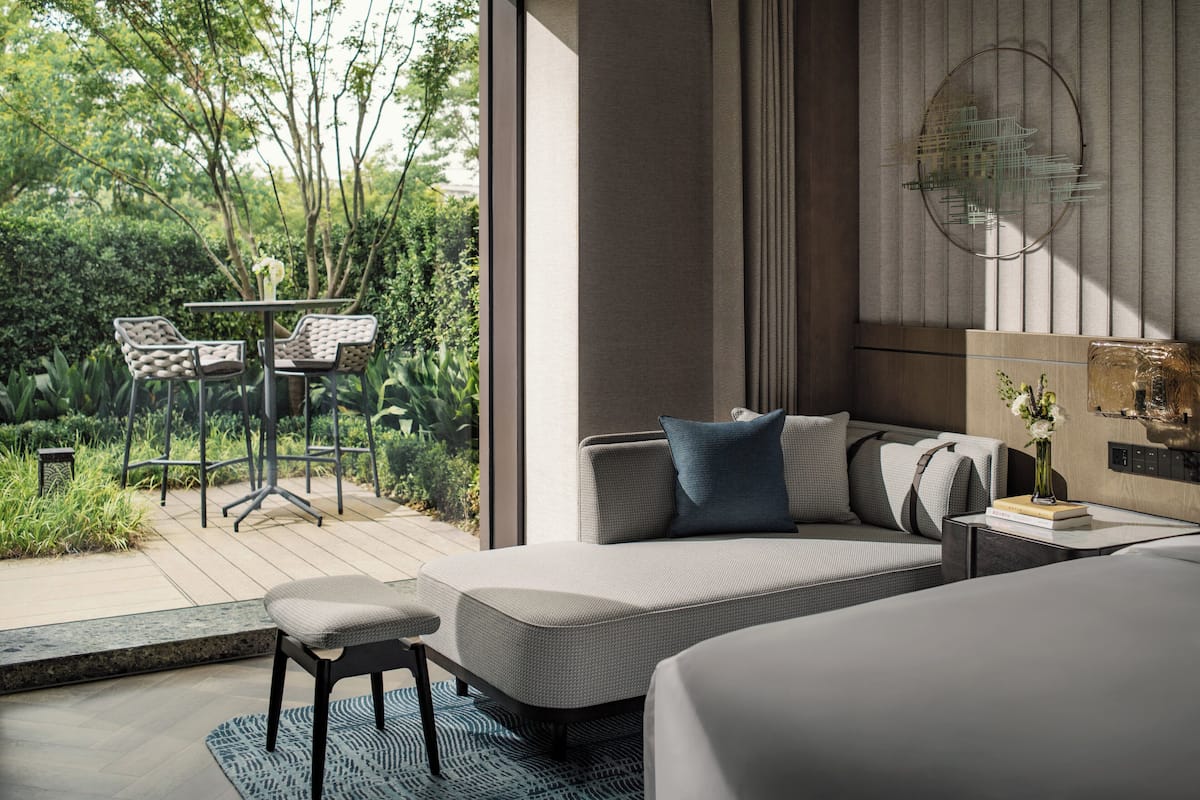 A modern bedroom with a large bed, a sofa, and a small table with chairs. There is a view of greenery outside the window.