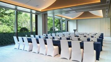 A conference room with rows of tables and chairs, large windows, and a view of greenery outside.