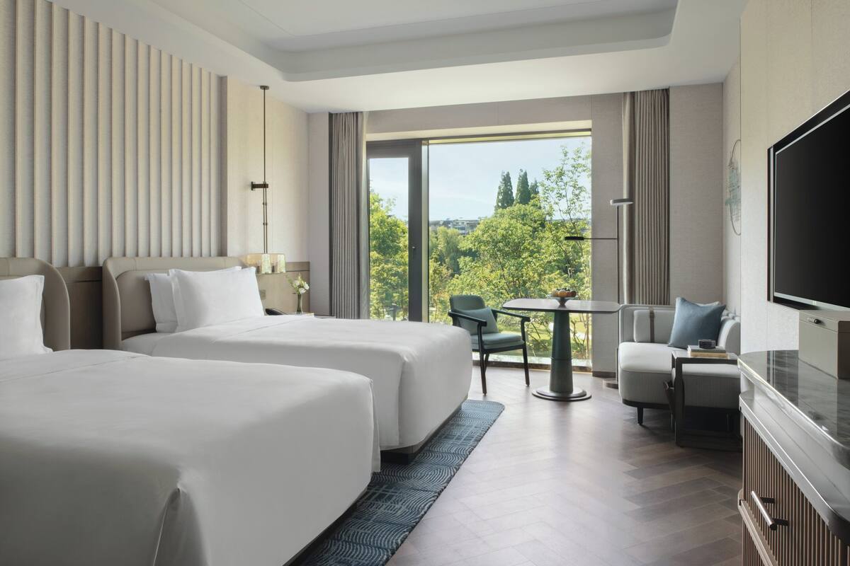 A hotel room with two beds, a television, a desk with a chair, and a view of trees and buildings.