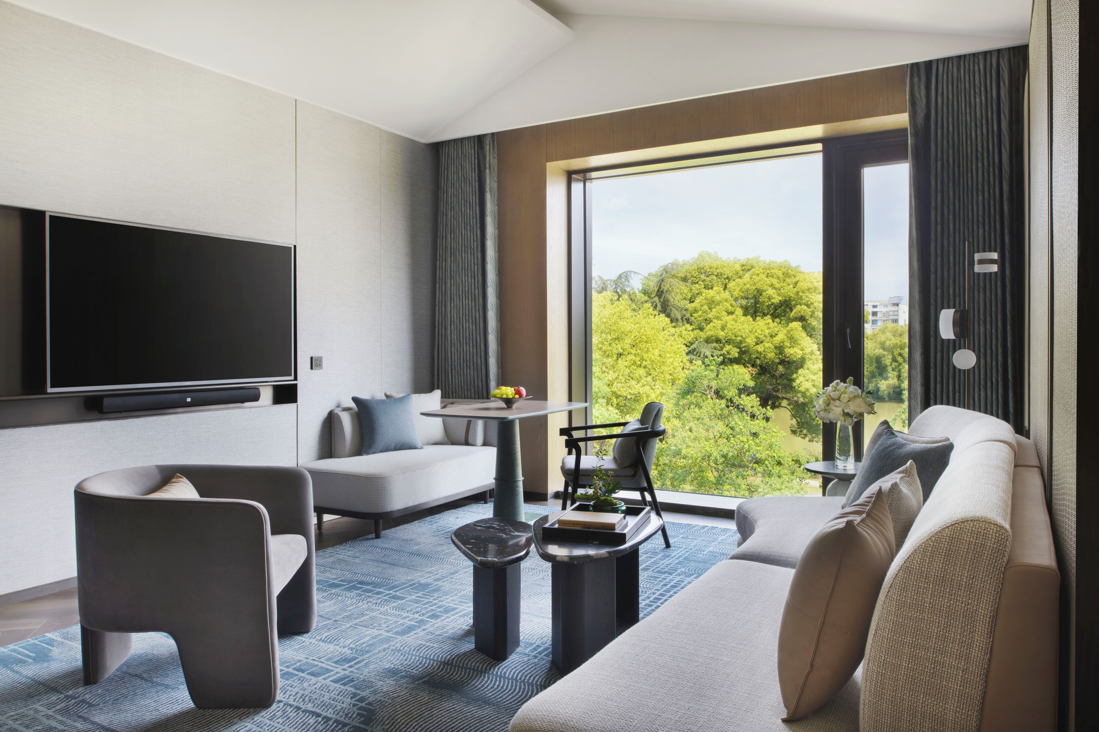 A modern living room with a flat-screen TV, a sofa, a coffee table, and a view of greenery through large windows.
