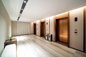 Corredor moderno com assoalho de madeira, portas de elevador numeradas e uma pequena mesa com uma planta.