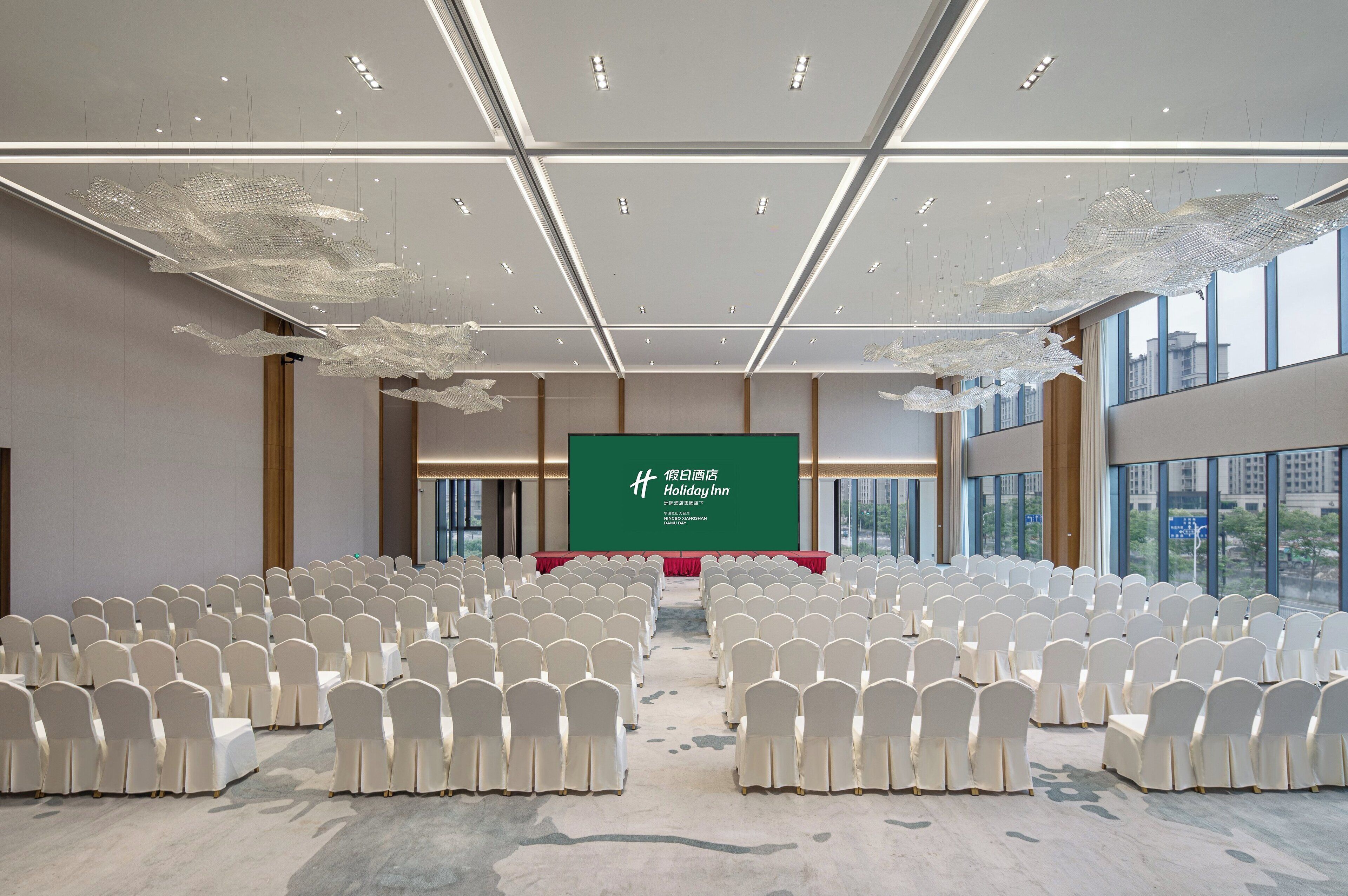 A conference room with rows of white chairs, a green screen with text, and large windows.