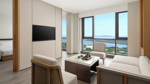 A modern living room with a large TV, comfortable seating, a coffee table with decorative items, and a view of the sea through large windows. - Holiday Inn Ningbo Xianshan Damu Bay by IHG (Ningbo)