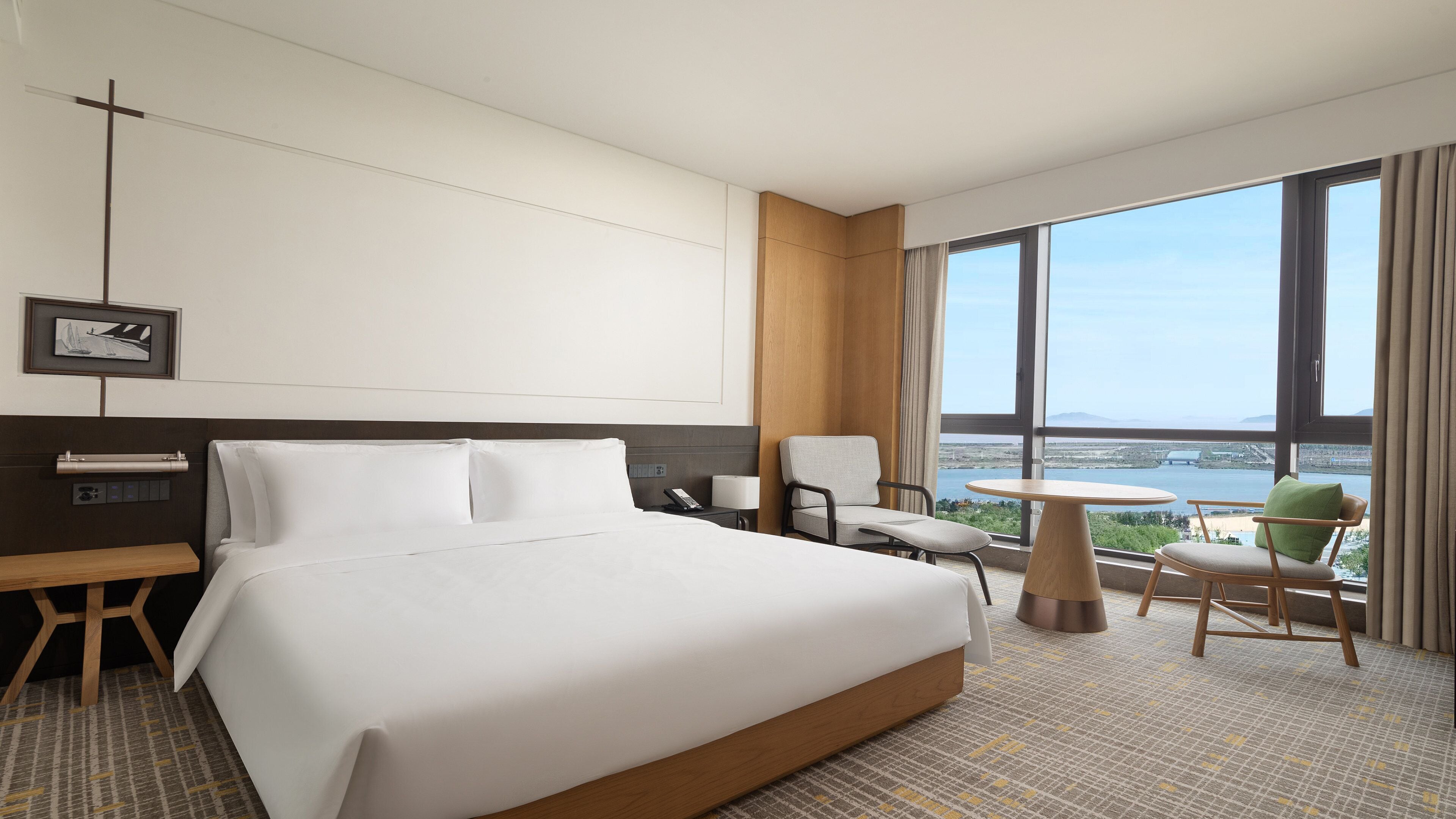 A hotel room with a large bed, a desk, a chair, a small table, and a view of a body of water through the window.