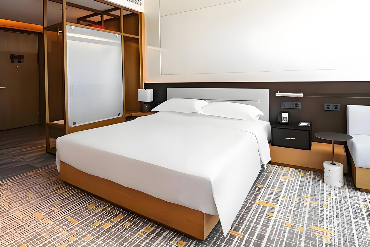 Quarto de hotel com uma cama grande, um guarda-roupa, uma mesa de cabeceira com abajur e uma pequena mesa com uma cadeira.