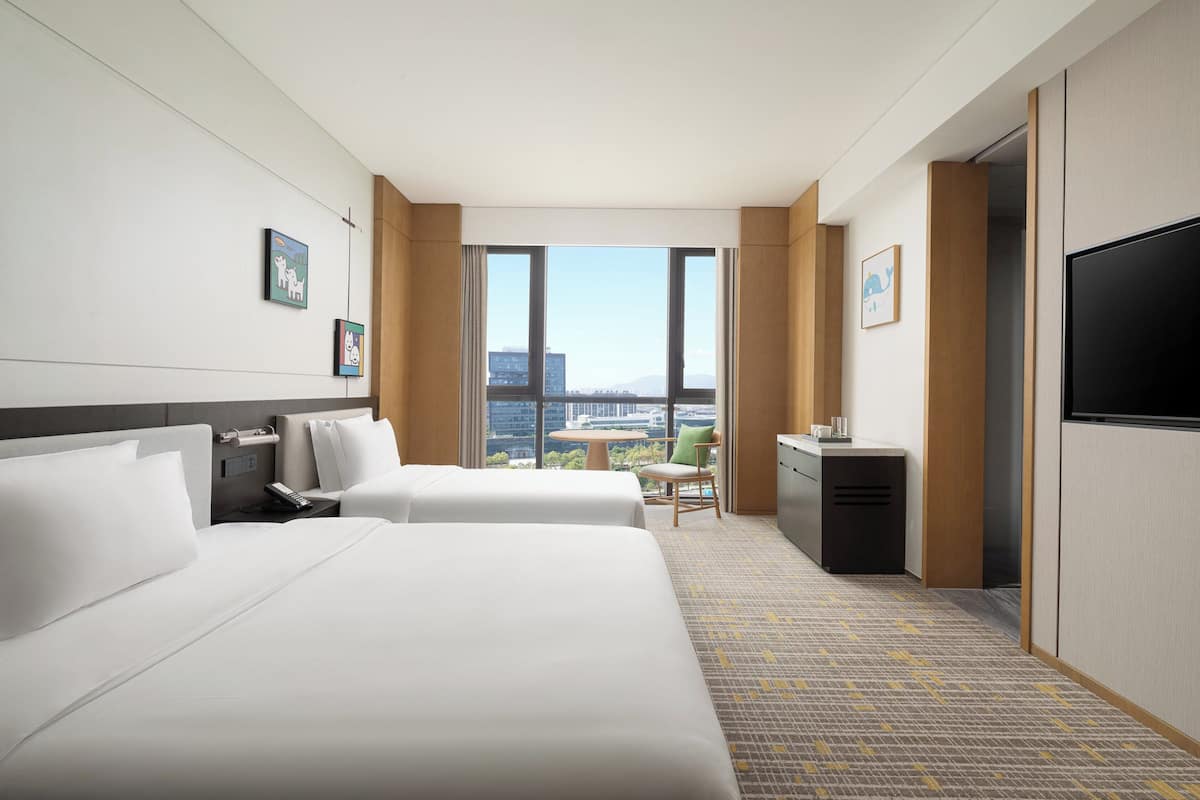 Een hotelkamer met twee bedden, een bureau, een stoel, een televisie en uitzicht op de stad.