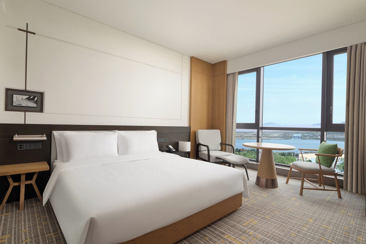 Quarto de hotel com uma cama grande, uma escrivaninha, uma cadeira, uma mesa pequena e vista para um corpo d'água através de amplas janelas.