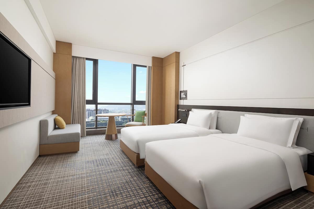 Hotelkamer met twee bedden, een bank, een televisie en een groot raam met uitzicht op de stad.