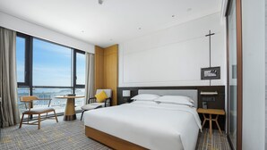 Quarto de hotel com uma cama grande, uma escrivaninha, uma cadeira, uma mesinha de centro e uma janela ampla com vista para a cidade.