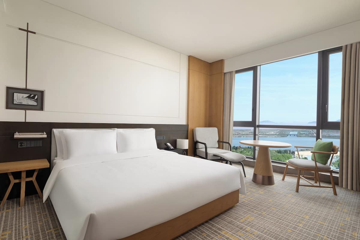 Hotelkamer met een groot bed, een bureau, een stoel, een tafeltje en uitzicht op een wateroppervlak door grote ramen.