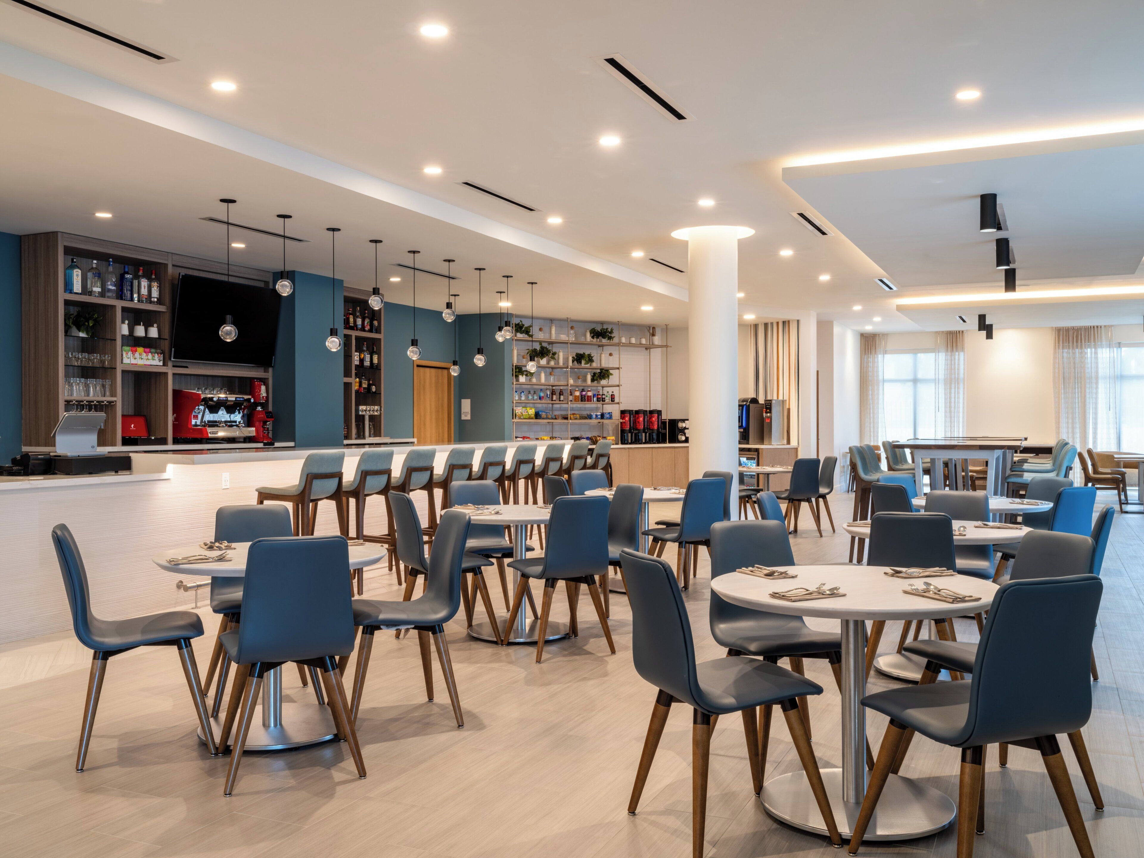 Un restaurante moderno con varias mesas y sillas, una zona de bar con una máquina de café y estanterías llenas de botellas y vasos.