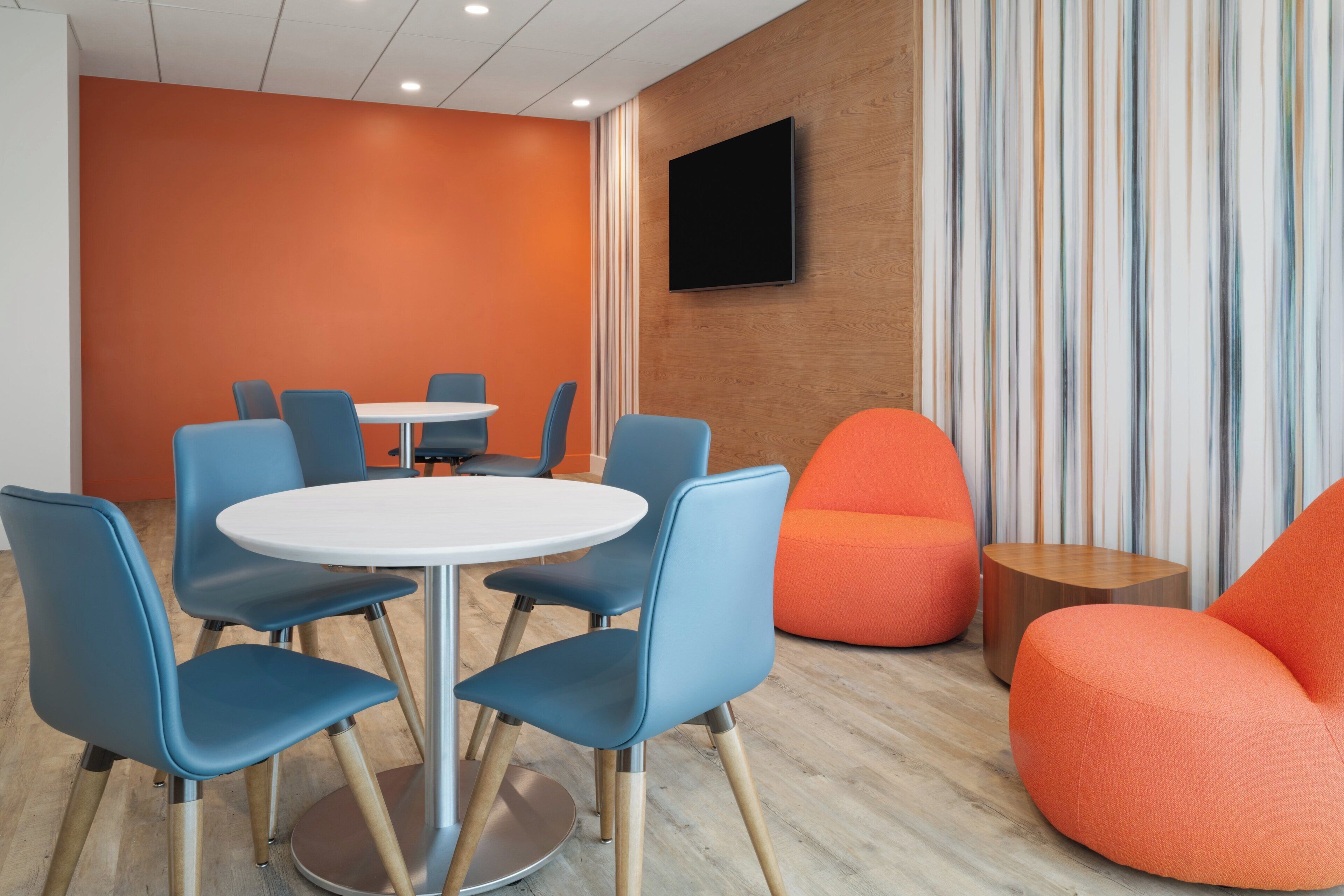 A modern meeting room with a white table and blue chairs, an orange sofa, and a TV on the wall.