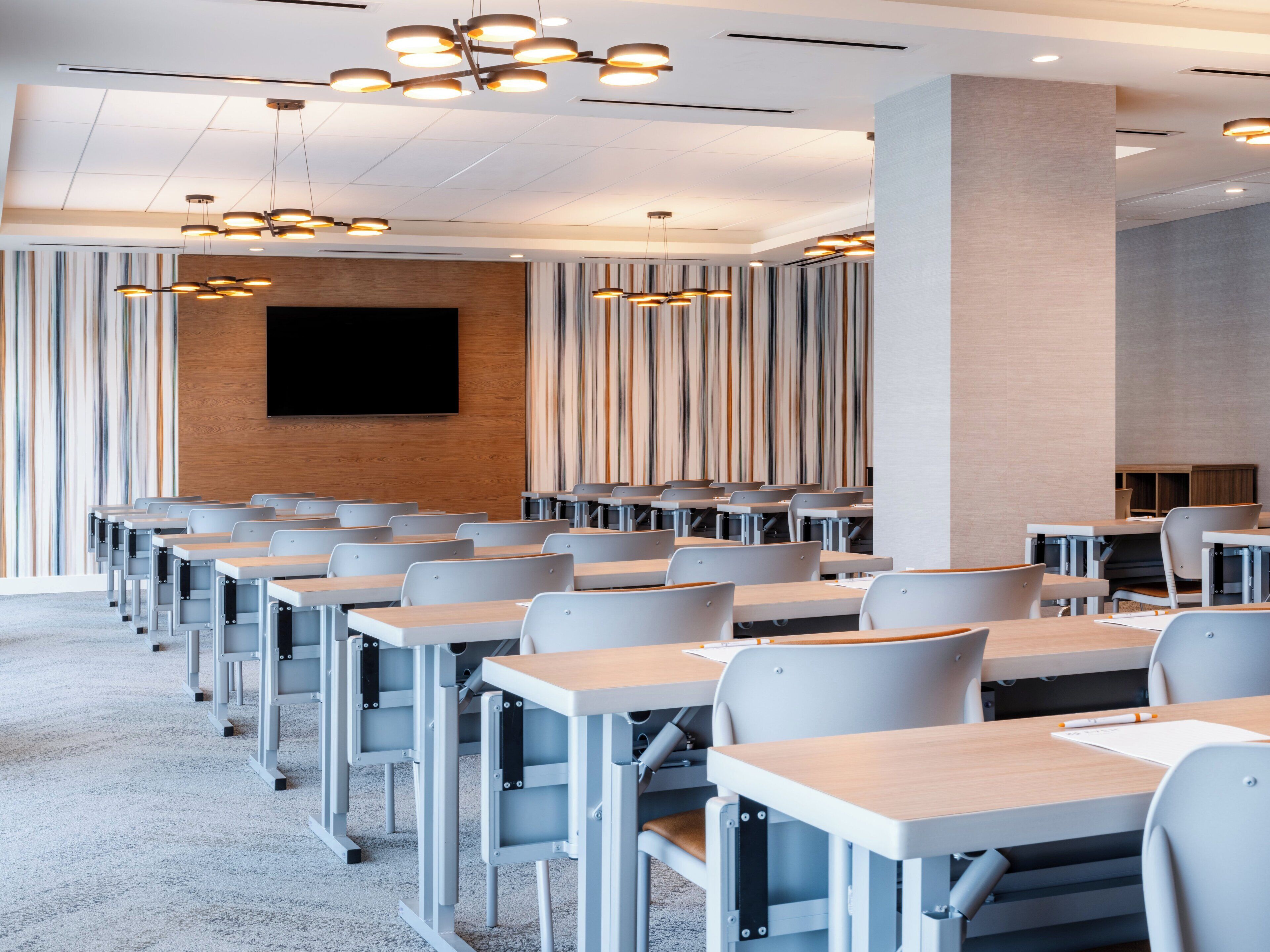 A modern conference room with rows of desks and chairs, a large flat-screen TV mounted on the wall, and contemporary lighting fixtures.