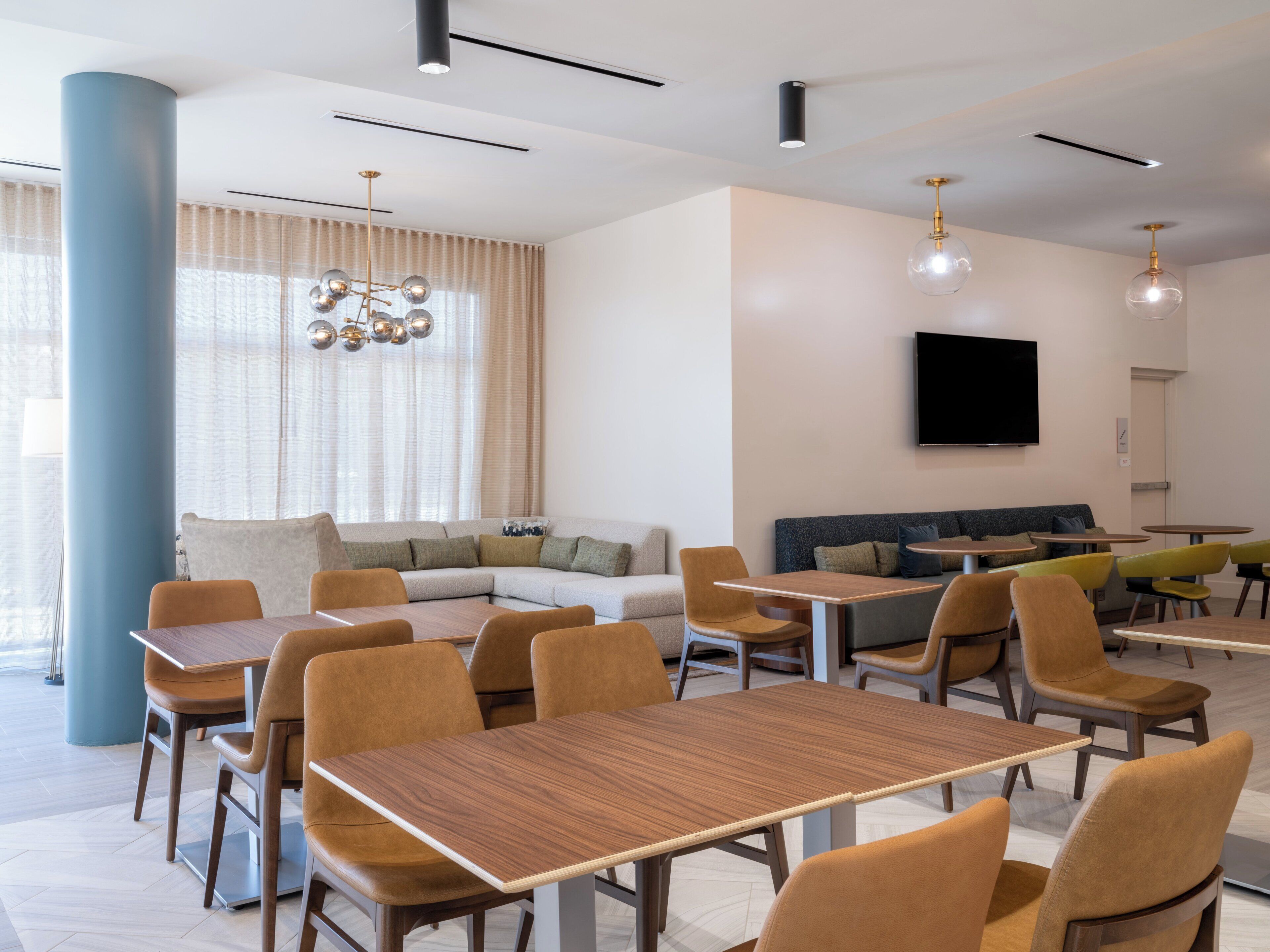 A modern dining area with multiple tables and chairs, a large window with curtains, a sofa, and a flat-screen TV mounted on the wall.