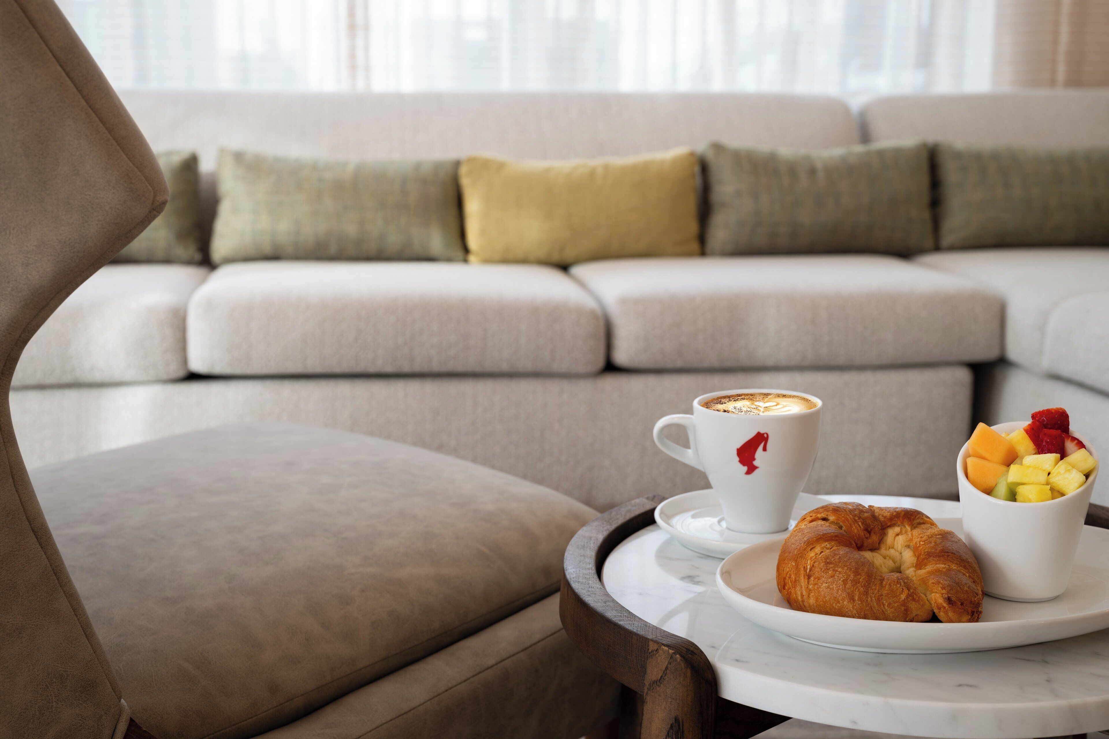 A cozy seating area with a cushioned sofa, a tray with a croissant and fruit, and a cup of coffee.