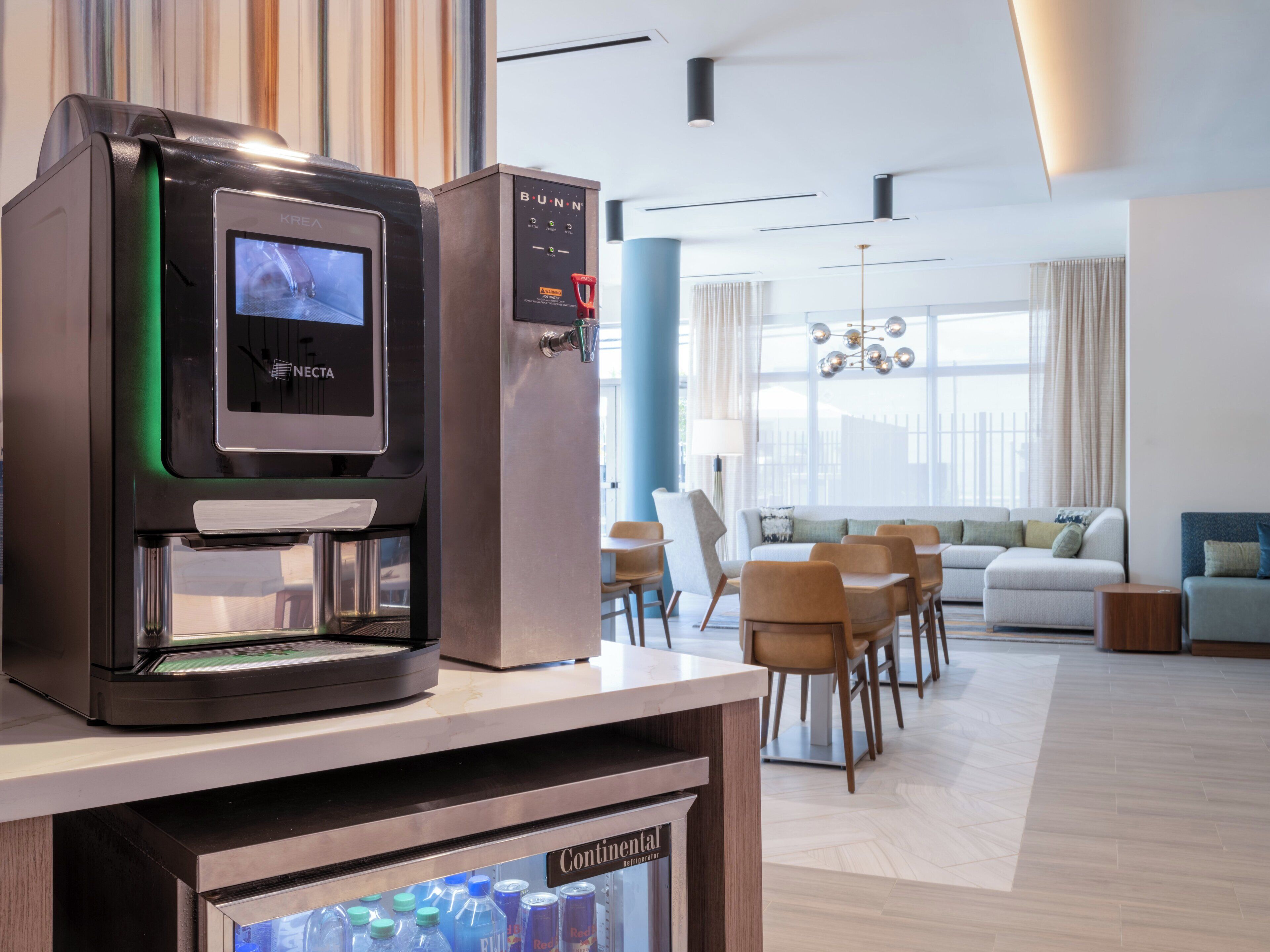 A modern kitchenette with a coffee machine, a mini-fridge stocked with beverages, and a seating area with a sofa and chairs.